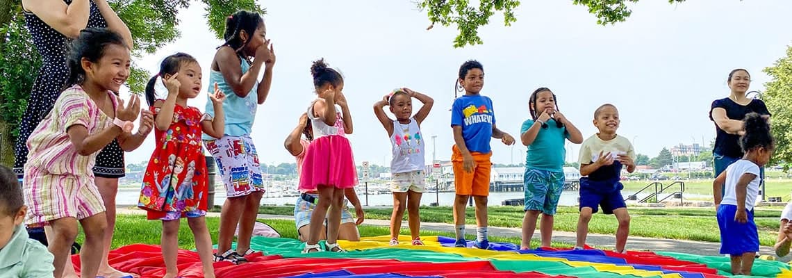 Children laughing and dancing on a rainbow parachute at an outdoor FNC playgroup by the waterfront