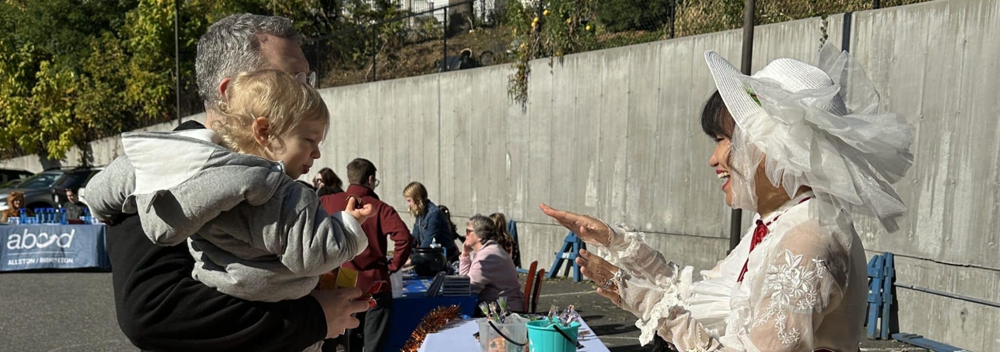 A parent holding a toddler greets a costumed performer at an outdoor fall festival hosted by FNC Allston-Brighton