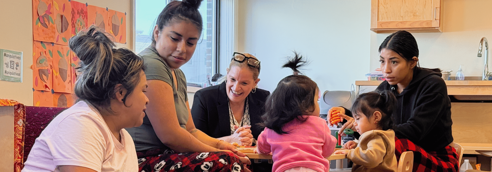Caregivers and toddlers playing together around an activity table in a classroom at FNC Dorchester