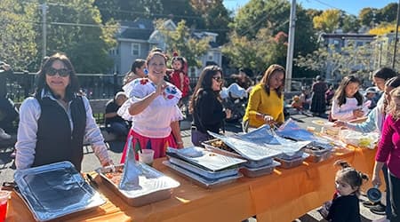 Volunteers serve food from a long table at an outdoor community celebration on a sunny fall day