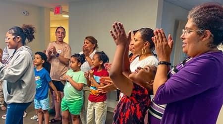 Caregivers and children stand together, clapping and singing, during a parenting education class