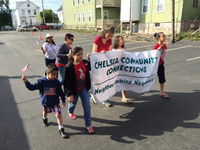 Children and caregivers march with the Chelsea Community Connections banner reading Neighbors Helping Neighbors