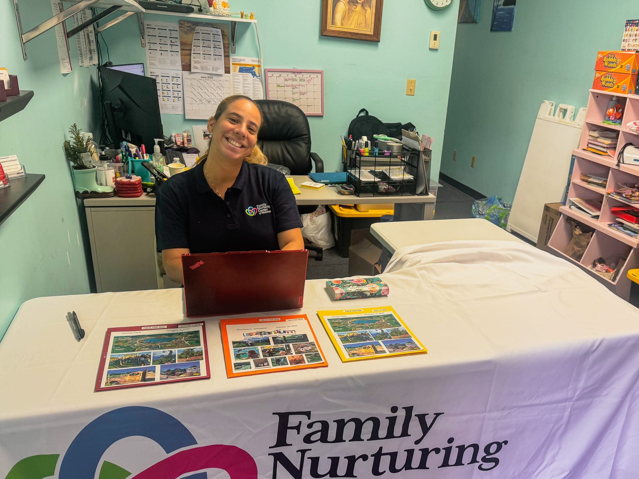 An FNC Chelsea staff member smiles at a welcome table displaying summer field trip flyers and a Family Nurturing Center banner
