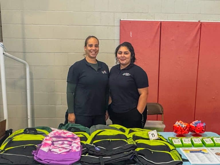 Two FNC Chelsea staff members stand behind tables of donated backpacks at a back-to-school giveaway