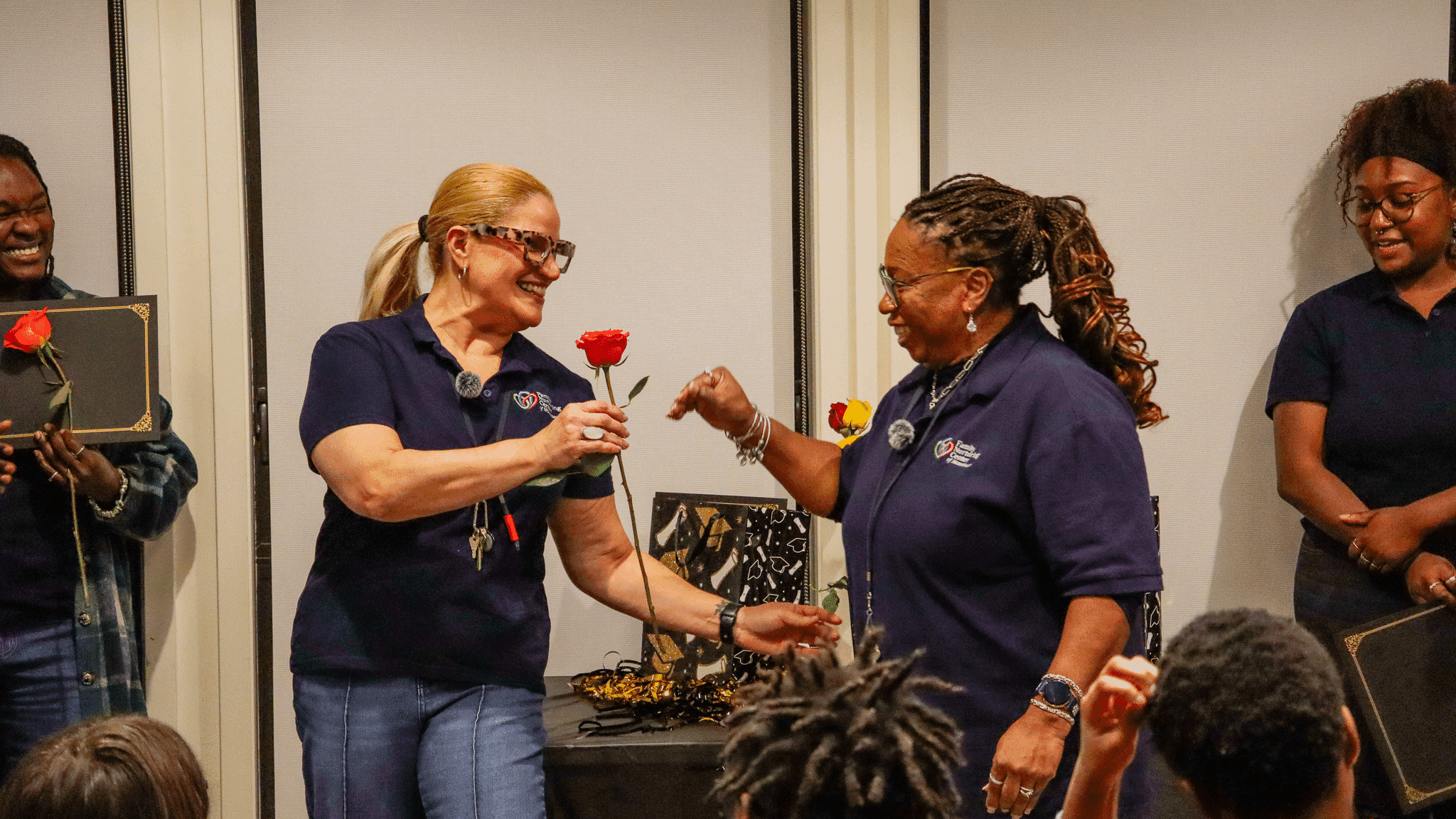 Two smiling Family Nurturing Center staff members in navy FNC polo shirts exchange roses at a team celebration