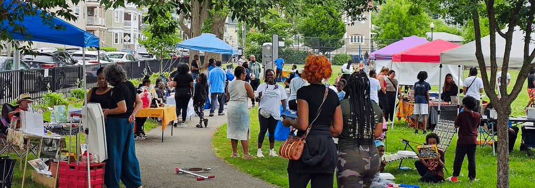 Neighbors of all ages browse tents and activity tables at an outdoor community festival in a city park.