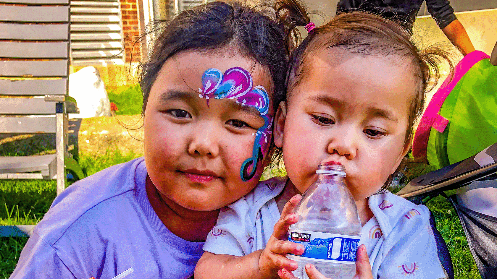 Two young children side by side at an outdoor celebration, one wearing butterfly face paint