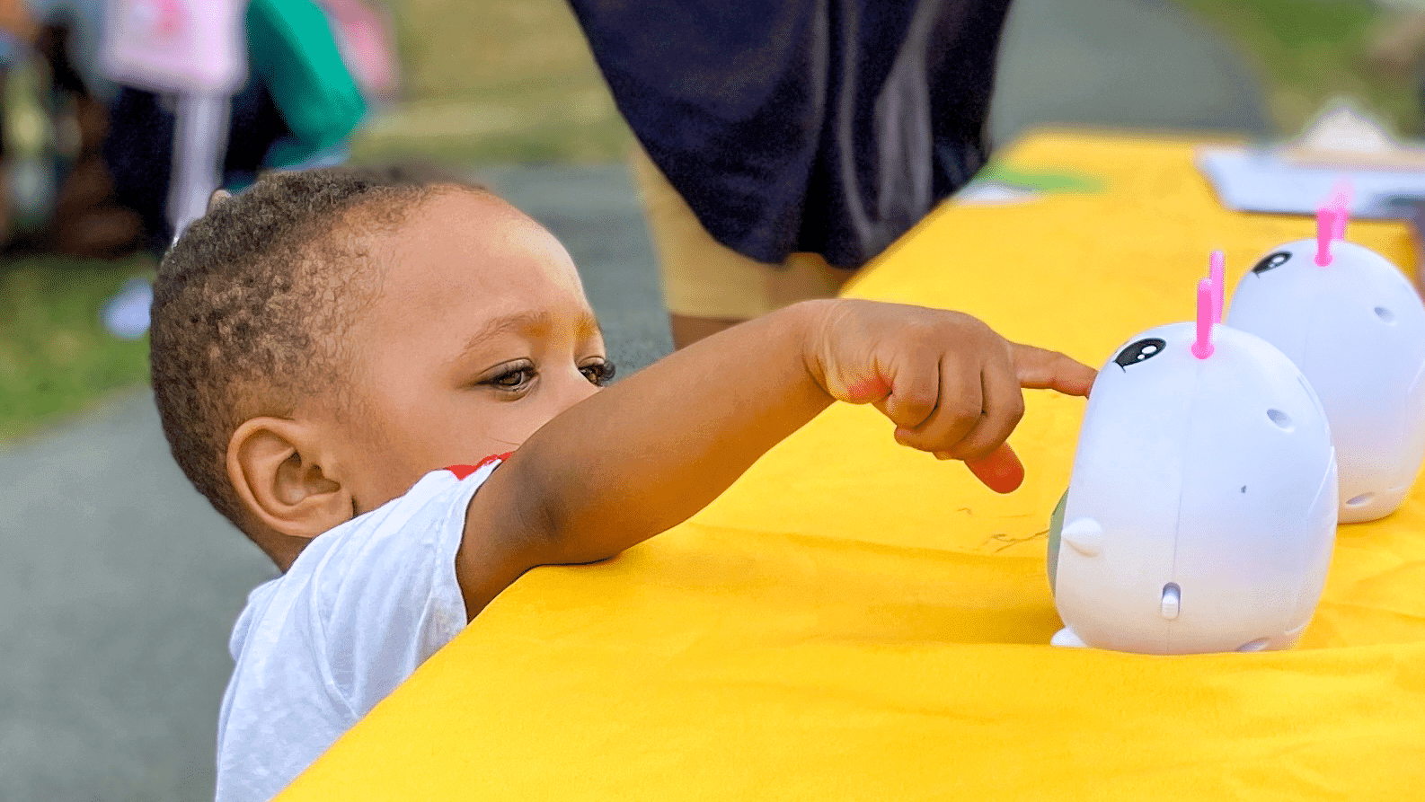 A toddler reaches across a yellow table to explore toys at an outdoor playgroup