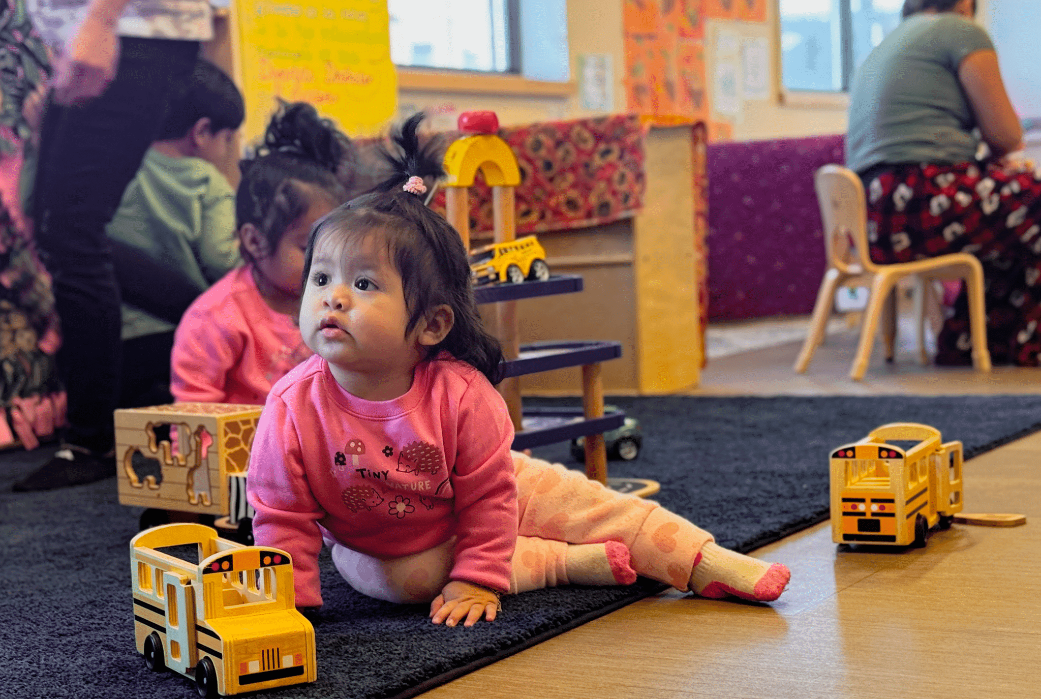 A baby crawls toward wooden toy school buses on a play mat while children and caregivers play together nearby.