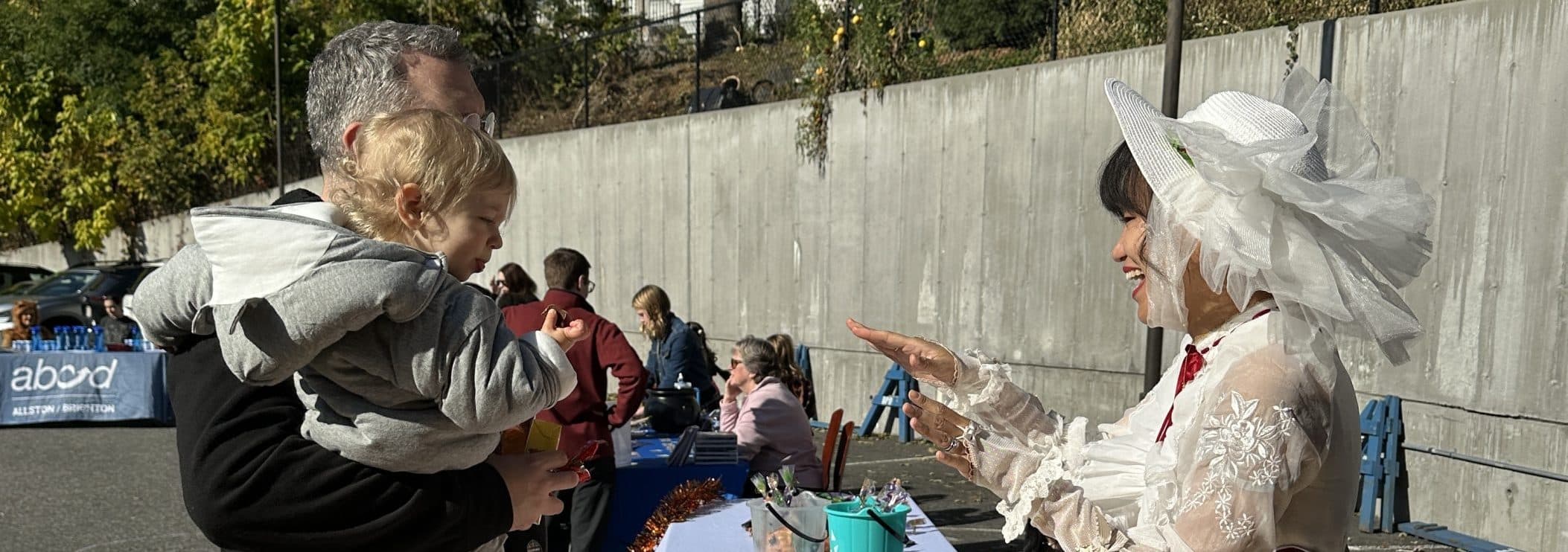 A parent holding a toddler greets a costumed performer at an outdoor FNC Allston-Brighton community event