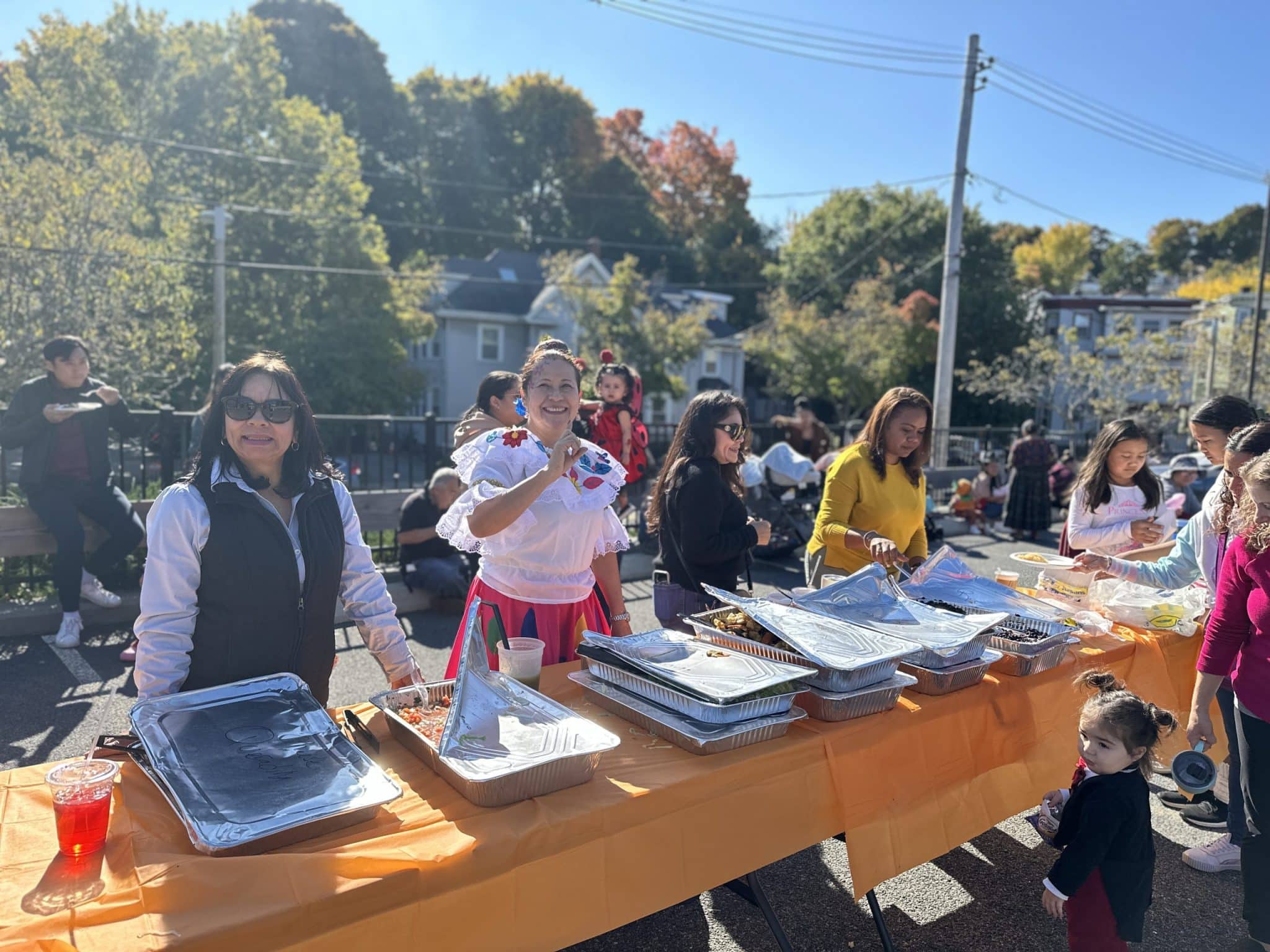 Neighbors and families share a meal at an outdoor community celebration on a sunny fall day, with a smiling woman in traditional dress serving food from a long table.