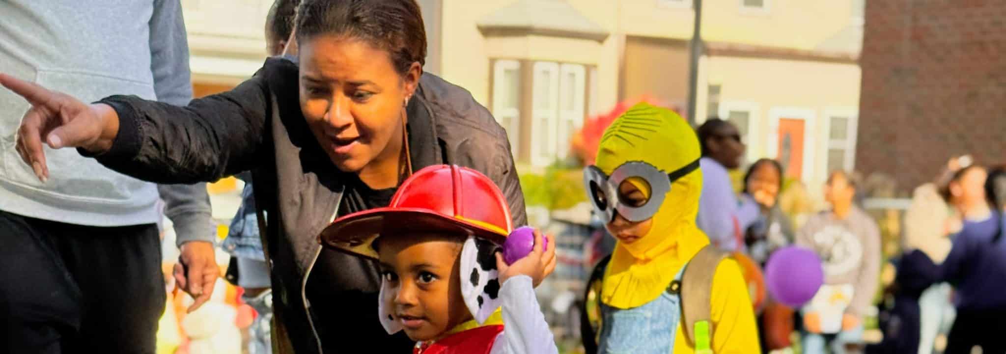 A caregiver points out an activity to two children in Halloween costumes at a neighborhood fall festival.