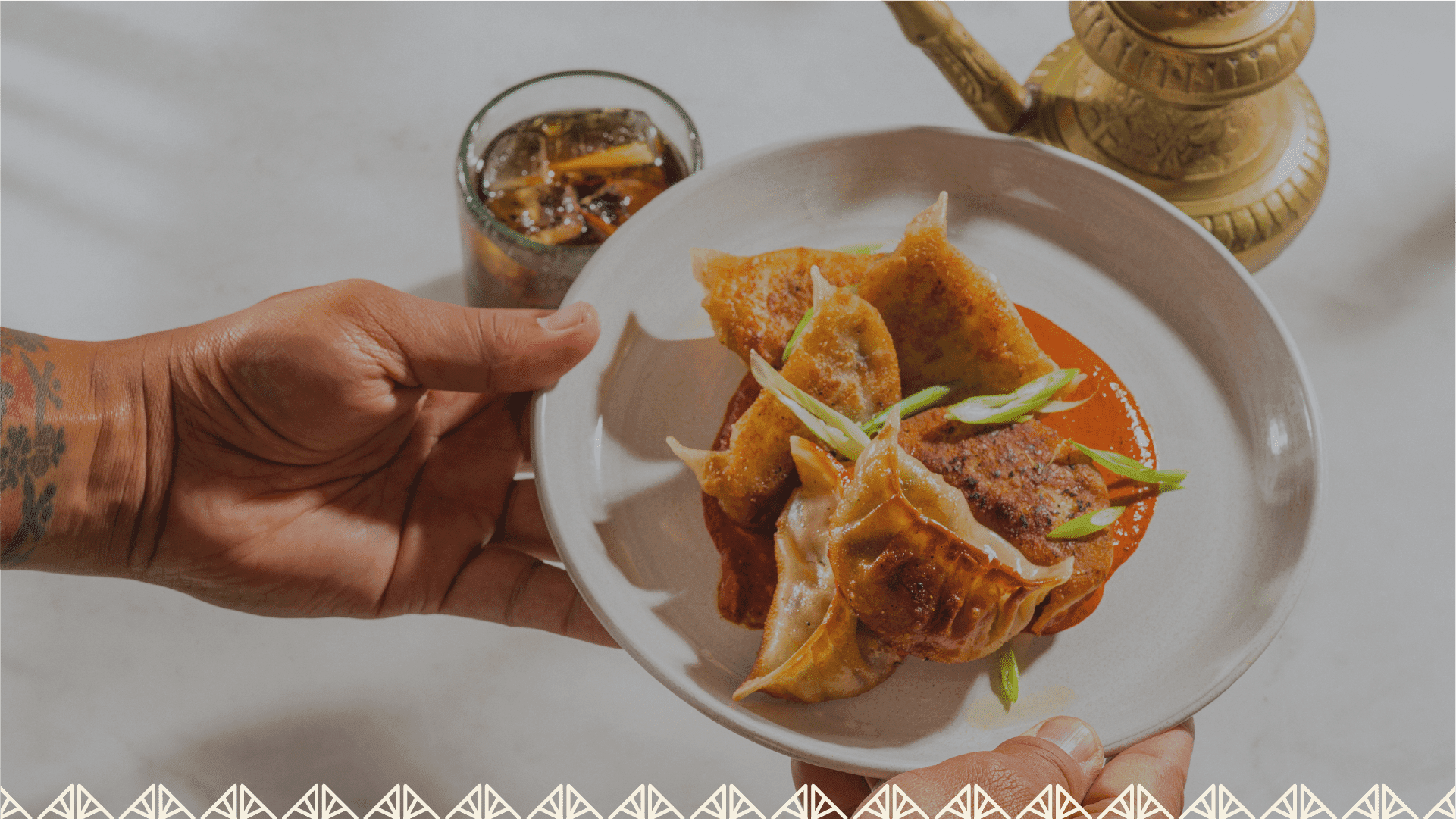 A server presents a plate of golden pan-seared dumplings over red sauce at Ama at The Atlas, beside an iced drink and a brass teapot