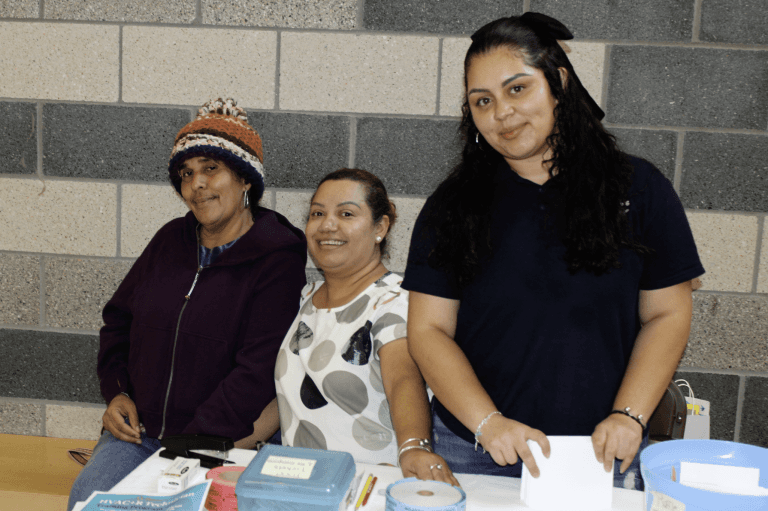 Three smiling women stand together behind a community resource table