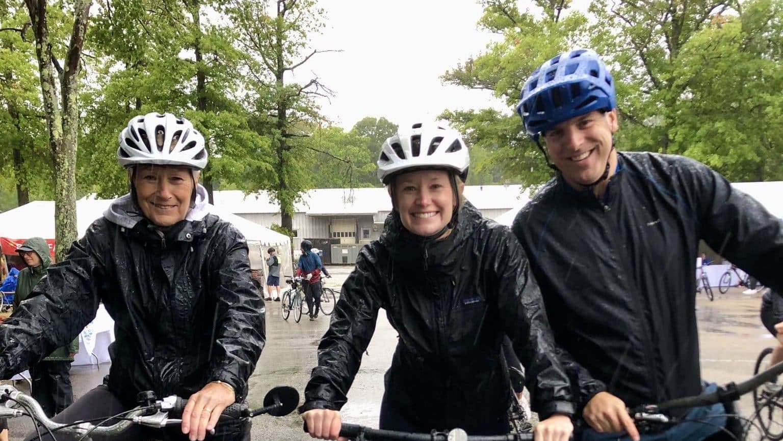 Three riders in rain jackets and helmets grin from their handlebars on a rainy morning