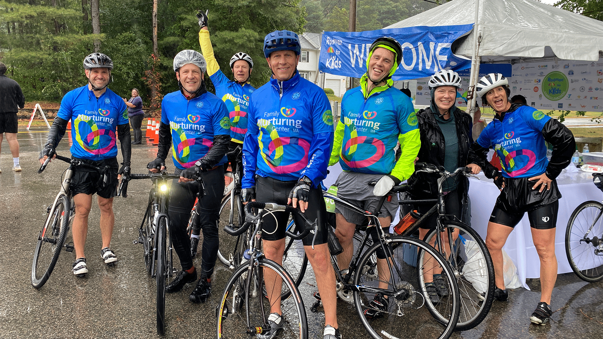 Seven Team FNC riders in Family Nurturing Center jerseys pose with their bikes by the welcome tent on a rainy Ride Day