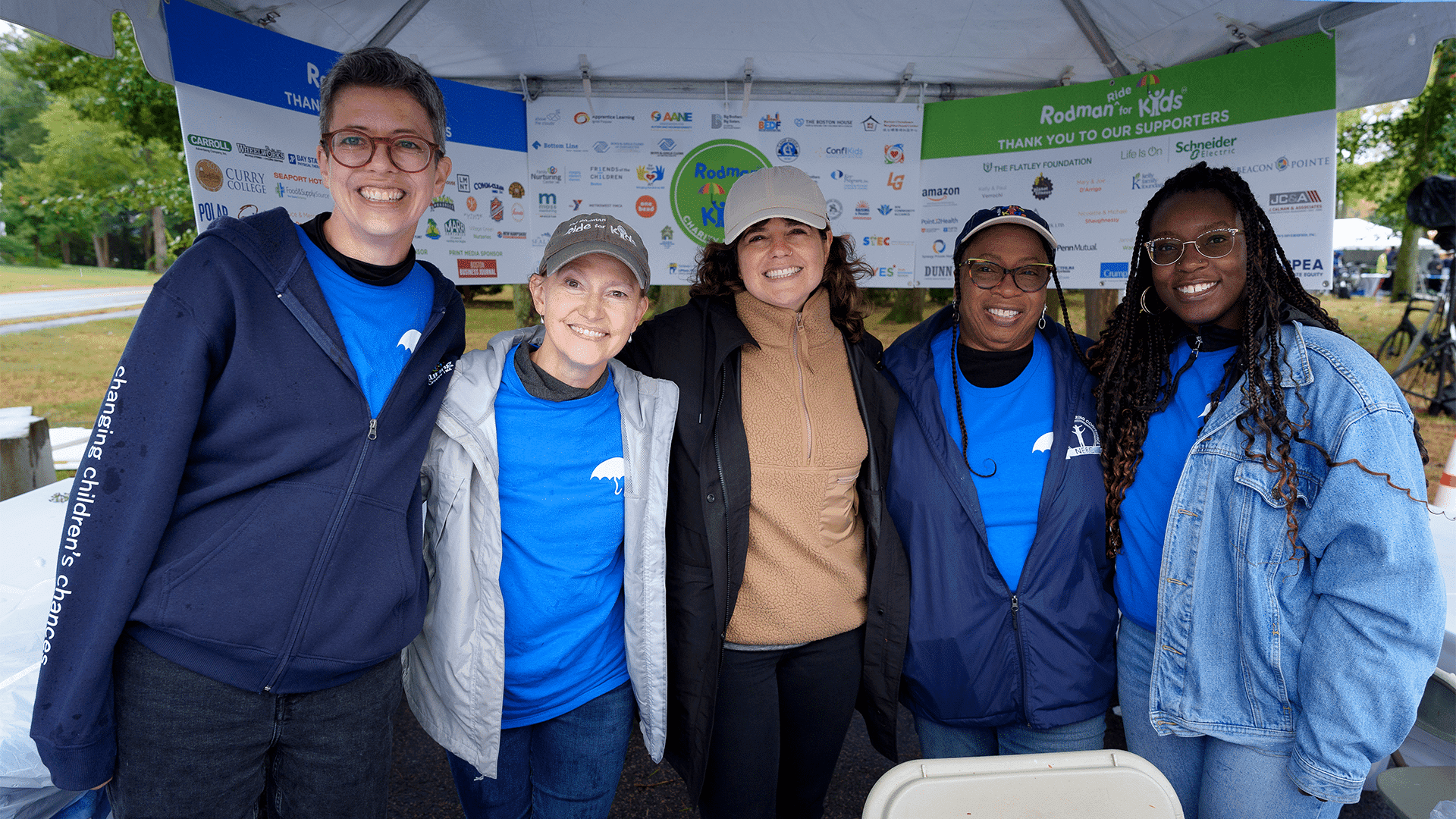 Five volunteers smile arm in arm under the Rodman for Kids supporters tent