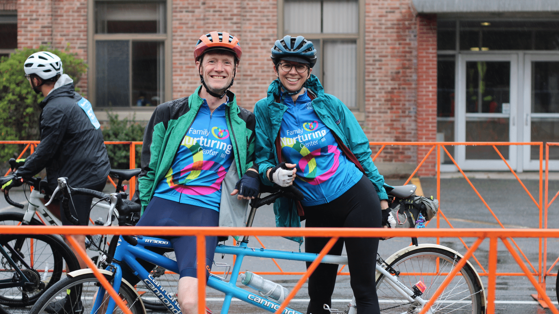 Two smiling riders in Family Nurturing Center jerseys stand with their tandem bike outside the ride venue