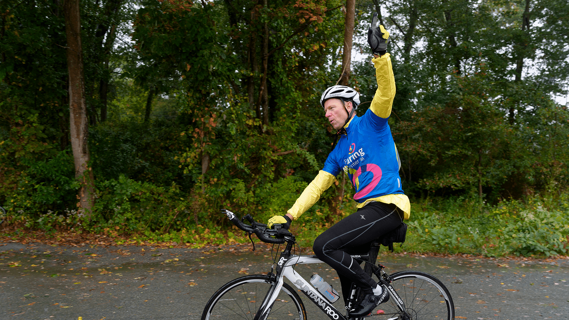 A Team FNC rider in a yellow rain jacket raises an arm in celebration while pedaling a rain-soaked road