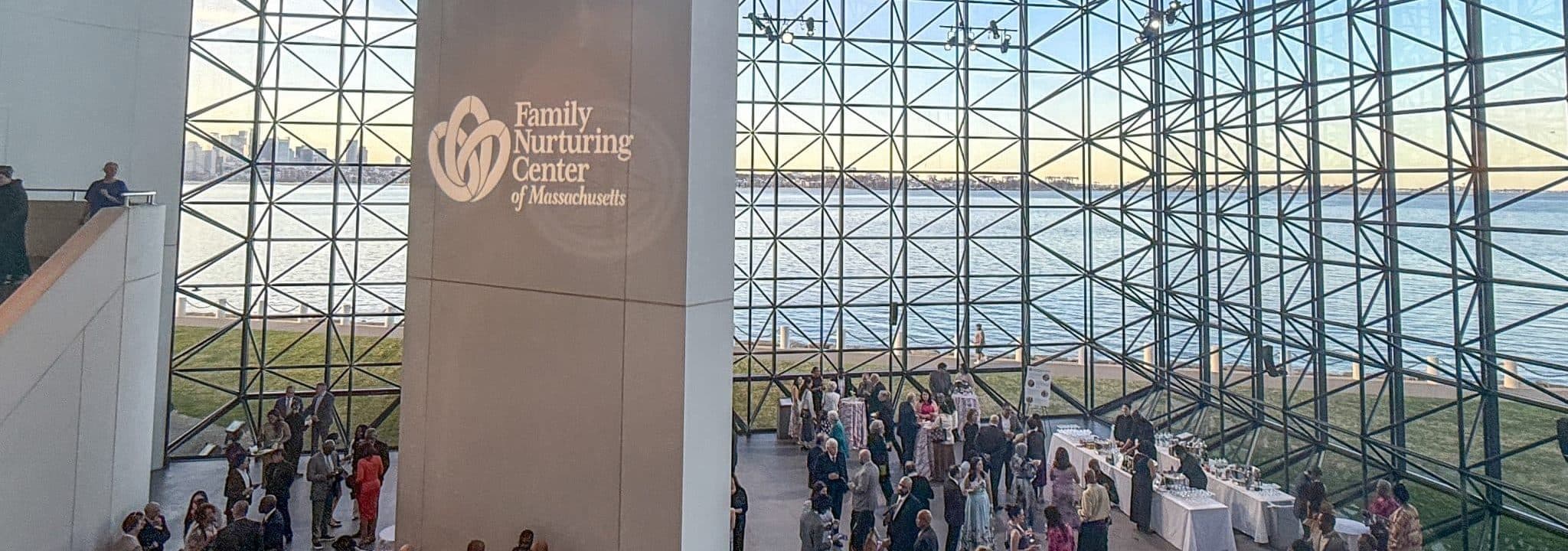 Gala guests mingle beneath the soaring glass wall of the JFK Presidential Library, with the FNC logo projected overhead and Boston Harbor beyond.